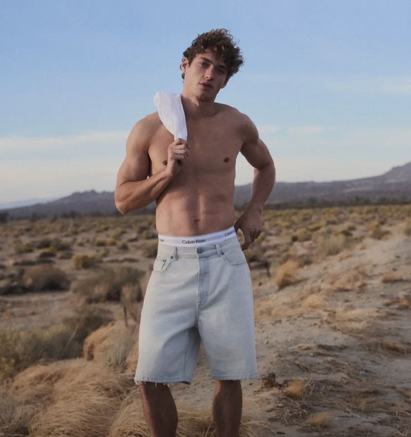 Shirtless young man standing outdoors in a desert landscape wearing light blue denim shorts with a white undergarment waistband visible