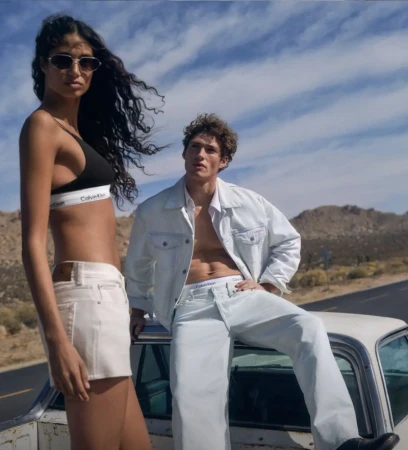 A tall woman and a man pose outdoors in desert landscape, wearing Calvin Klein branded denim and underwear. The photo is taken from low angle.