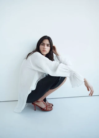 Model Loli Bahia squatting in a white blouse and dark trousers against a minimalist plain white background.