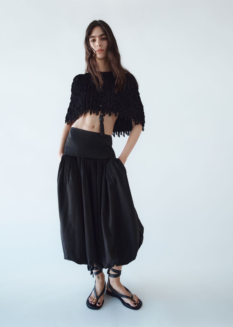 Model Loli Bahia in a black fringed crop top and loose skirt against a bright white background