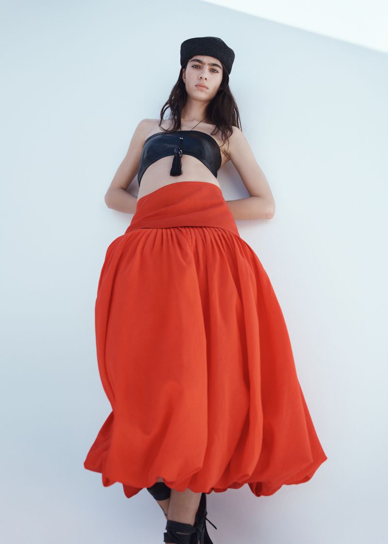Model Loli Bahia wearing a black bandeau top and long red skirt against a white studio background for H&M S/S 2026