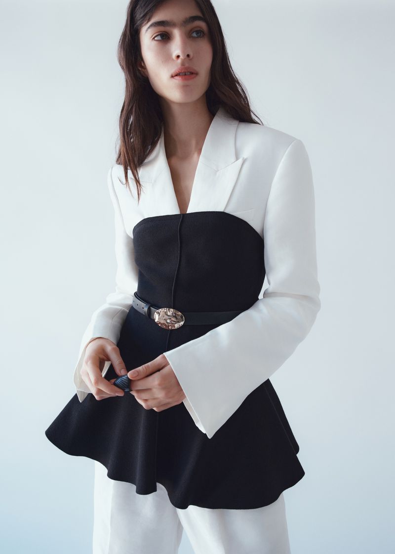 Model Loli Bahia wearing a black and white tailored garment against a light grey studio background.