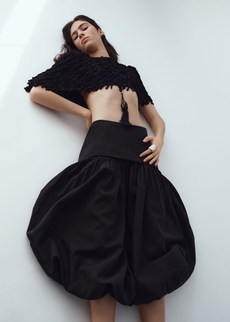Model Loli Bahia wearing a black textured crop top and voluminous dark skirt against a plain white background for H and M