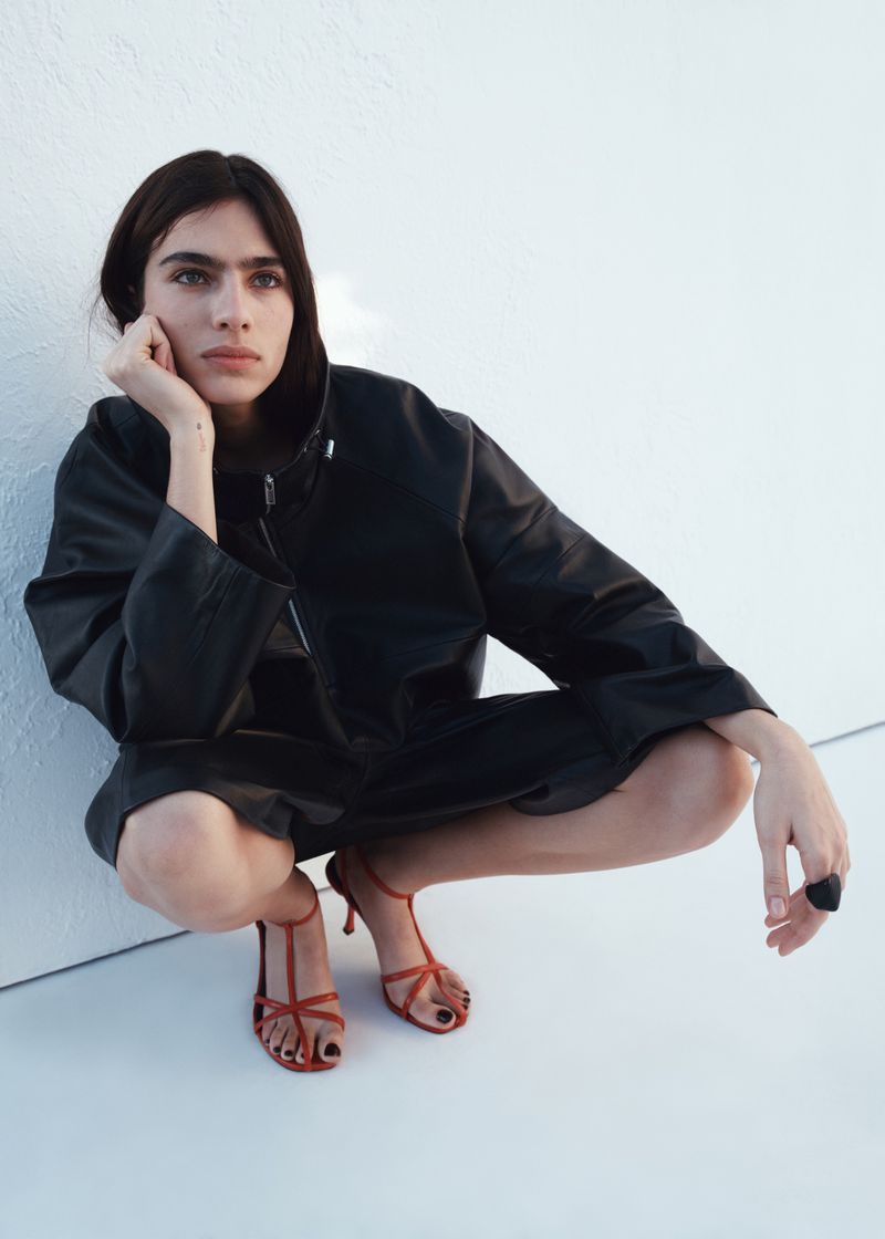 Model Loli Bahia crouching against a white background wearing a black jacket and red strappy heels for H&M S/S 2026