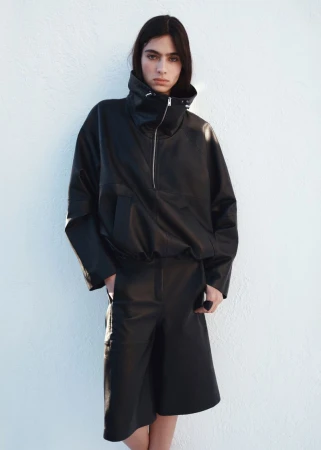 Model wearing a black oversized high-neck jacket and matching shorts against a white textured wall