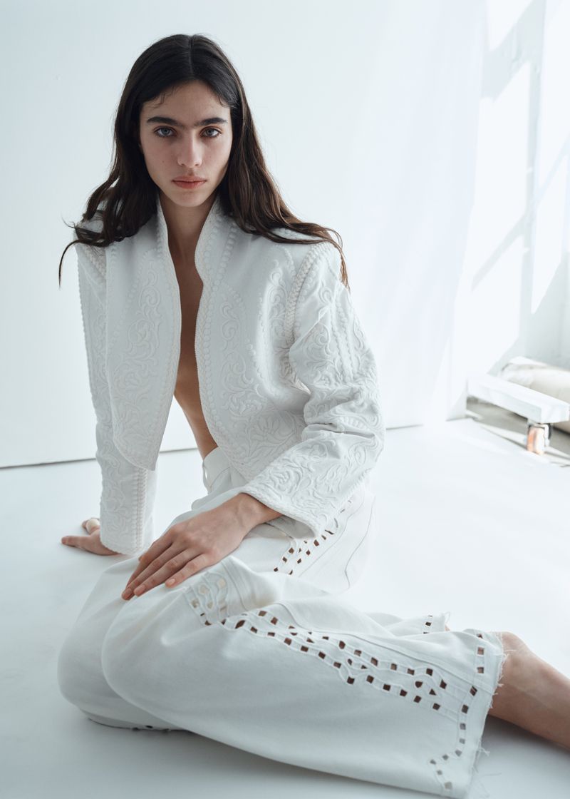 Model Loli Bahia seated on a white floor wearing an embroidered white jacket and matching cutout trousers for H&M.