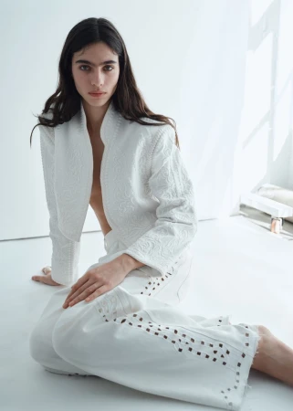 Model Loli Bahia seated on a white floor wearing an embroidered white jacket and matching cutout trousers for H&M.