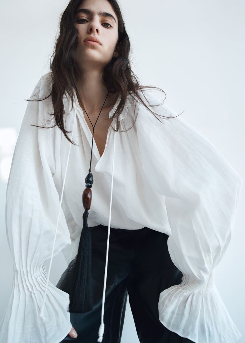 Dark-haired woman in a white blouse with a long tassel necklace poses in front of a white background in a fashion or editorial shot.