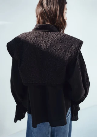 Model seen from behind wearing an embroidered black jacket over blue jeans against a pale backdrop