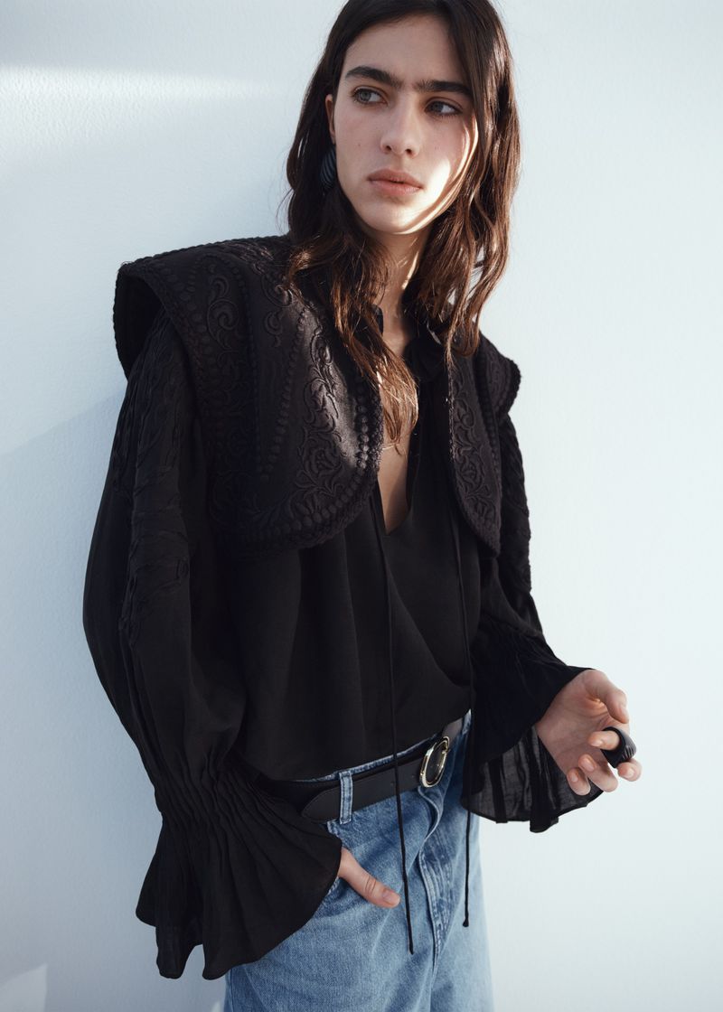Model Loli Bahia wearing a black embroidered blouse and denim jeans against a white seamless wall.