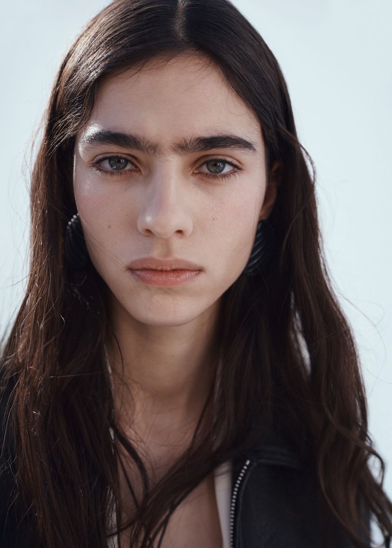 Close-up portrait of model Loli Bahia with long dark hair wearing a black jacket against a plain white background.