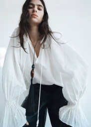 Model wearing a white blouse with voluminous sleeves and a tassel necklace against a white background