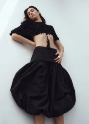 Model Loli Bahia wearing a black textured crop top and voluminous dark skirt against a plain white background for H and M