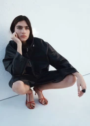 Model Loli Bahia crouching against a white background wearing a black jacket and red strappy heels for H&M S/S 2026