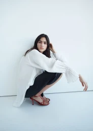 Model Loli Bahia squatting in a white blouse and dark trousers against a minimalist plain white background.