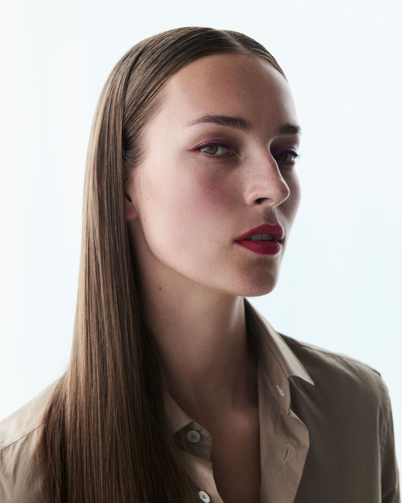 Close-up portrait of a woman with long straight brown hair wearing a beige button-down shirt and vibrant red lipstick against a bright white background