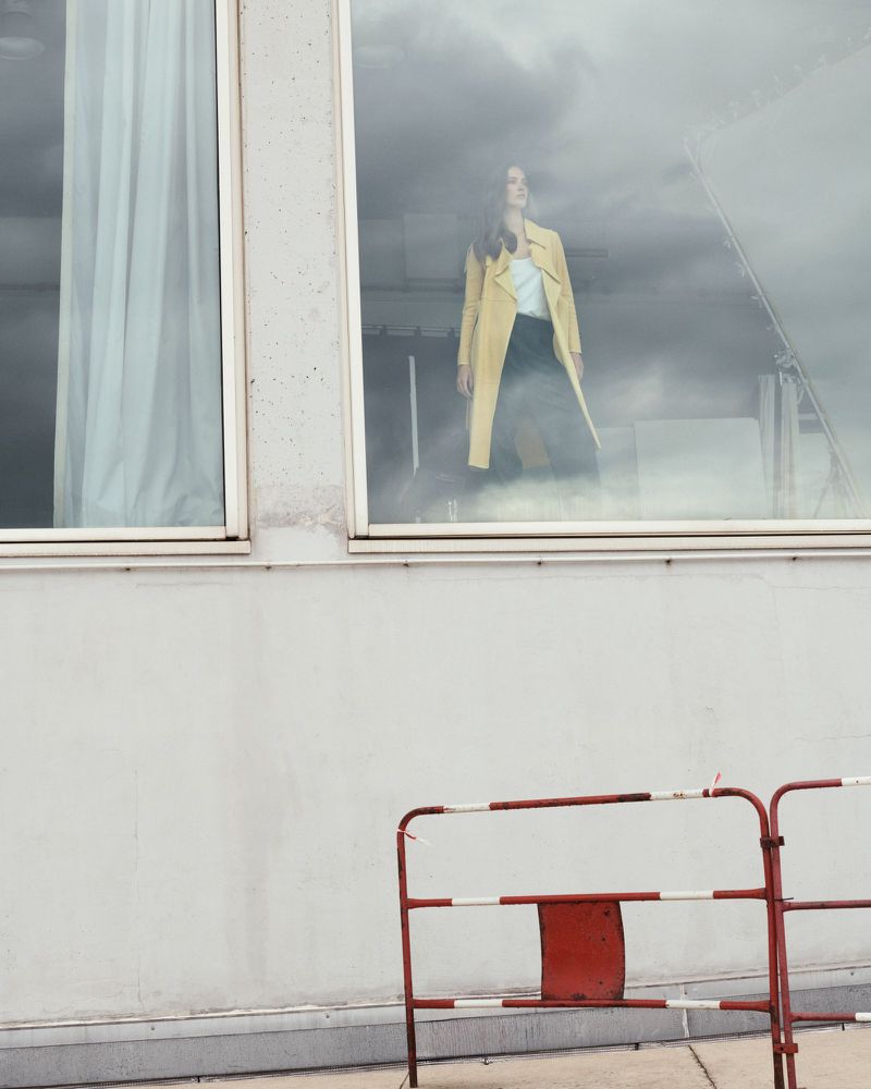 Fashion shot through a window: A woman in a yellow coat stands inside a building, seen through a window with a red barrier outside.