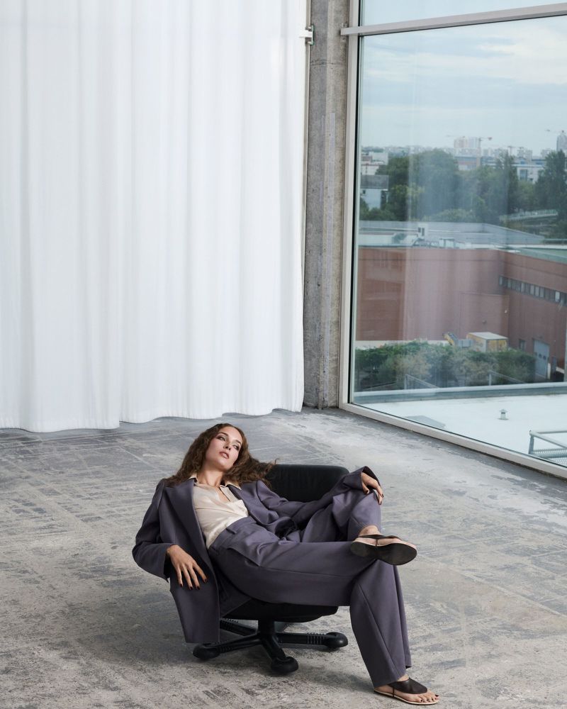 Woman in a grey suit seated in an office chair against a large window and white curtain background