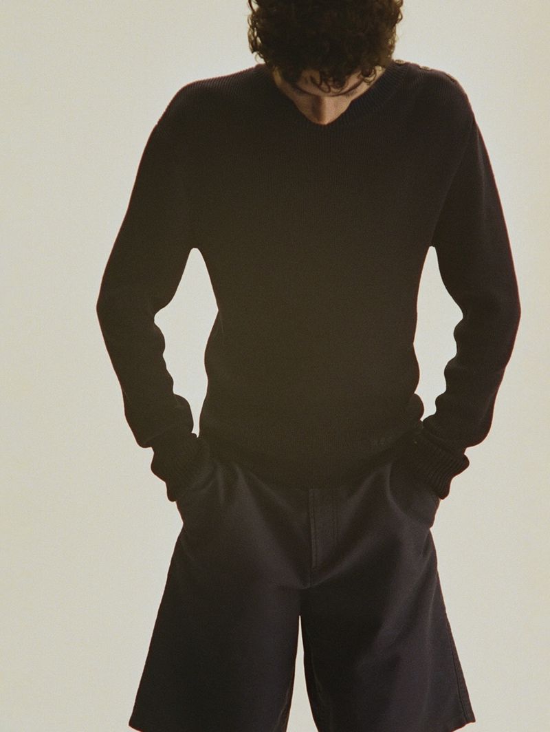 An adult male is posing indoors wearing a dark v-neck sweater and short shorts, with his hands in his pockets, against a plain backdrop.