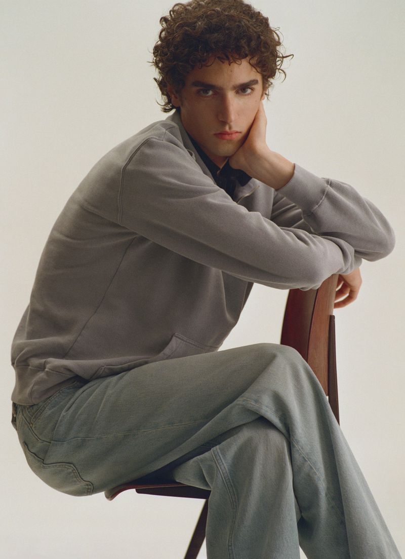Young man with curly hair seated backwards on a wooden chair wearing a grey sweatshirt and denim trousers against a white backdrop