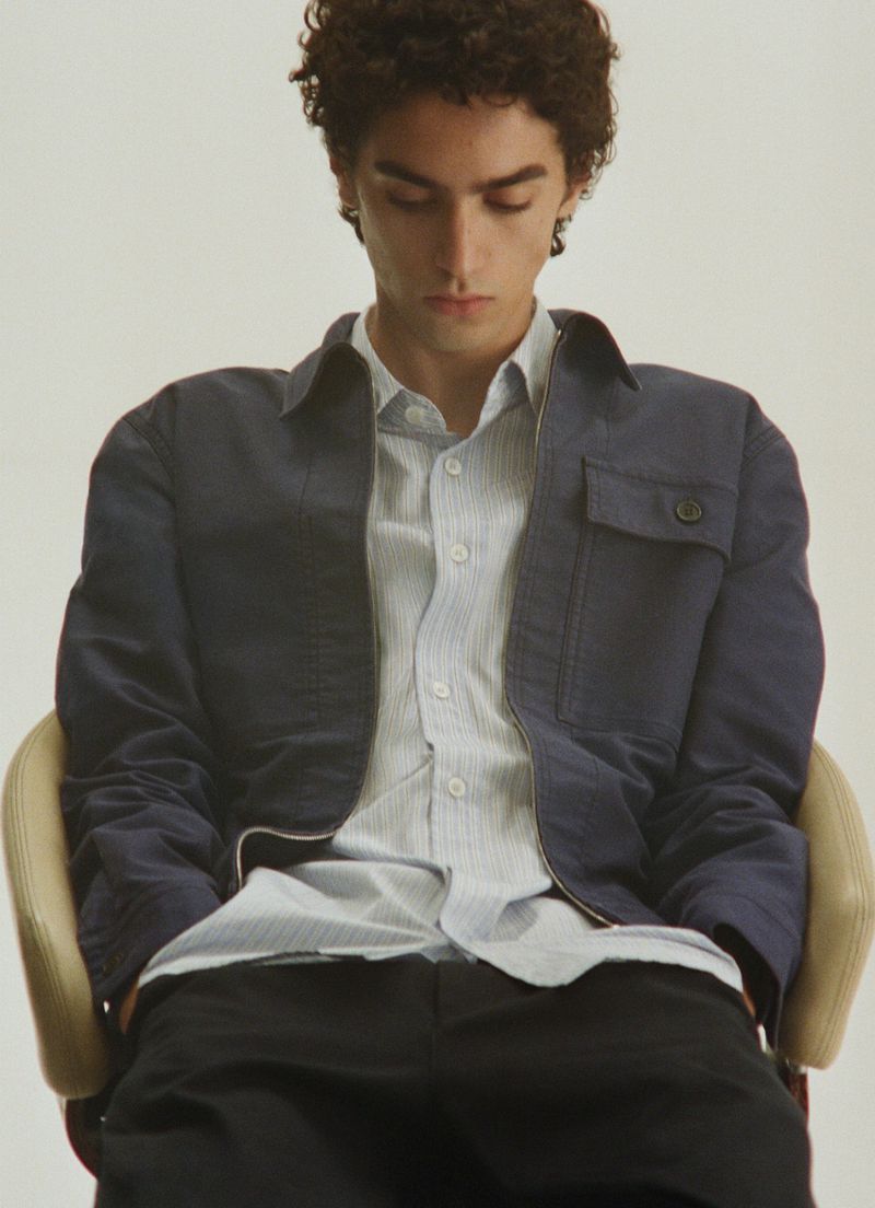 Young male model with curly dark hair seated in a chair wearing a navy jacket and striped button-down shirt.
