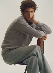 Young man with curly hair seated backwards on a wooden chair wearing a grey sweatshirt and denim trousers against a white backdrop