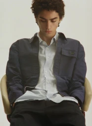 Young male model with curly dark hair seated in a chair wearing a navy jacket and striped button-down shirt.