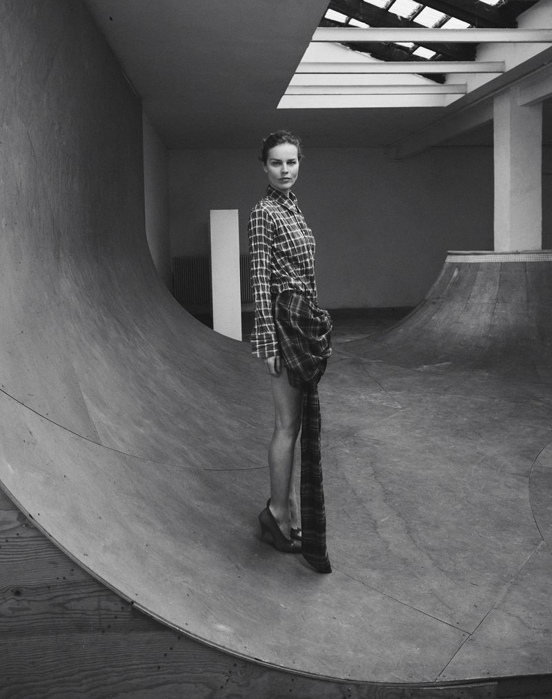 Monochrome fashion shot of a young woman in a plaid shirt dress posing in a concrete skatepark with a skylight.
