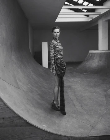 Monochrome fashion shot of a young woman in a plaid shirt dress posing in a concrete skatepark with a skylight.
