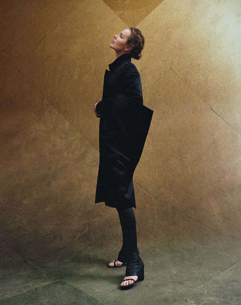 Adult female in a dark coat and sandals poses in a full shot against a tan and brown backdrop. She looks upward with neutral expression.