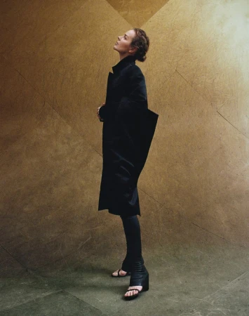 Adult female in a dark coat and sandals poses in a full shot against a tan and brown backdrop. She looks upward with neutral expression.