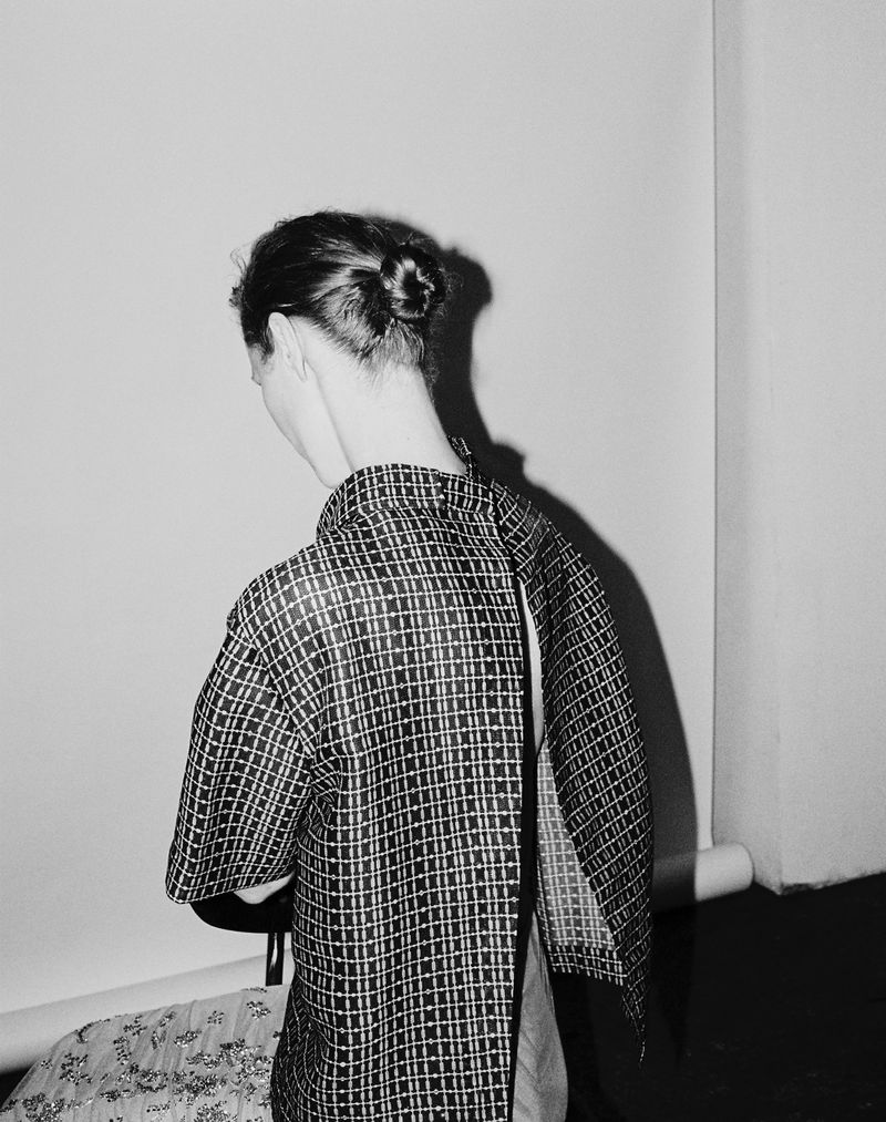 Back view of a woman with a bun wearing a patterned top against a white wall, creating a simple, monochrome fashion aesthetic.