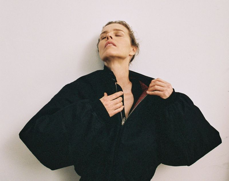 Adult female in black puffed-sleeve jacket, unzipped, eyes closed, head tilted back slightly, against a white wall, indoor studio shot.