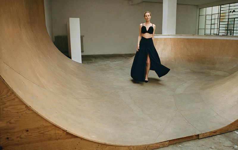Fashion editorial shot of a female model in a black bra and skirt, posing in a skatepark with a light-toned concrete and wood bowl.