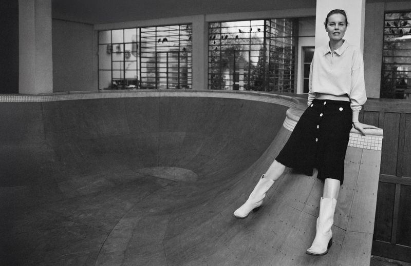 Monochrome studio shot of a woman in a black skirt and white boots sitting on the edge of a concrete halfpipe, architectural background.
