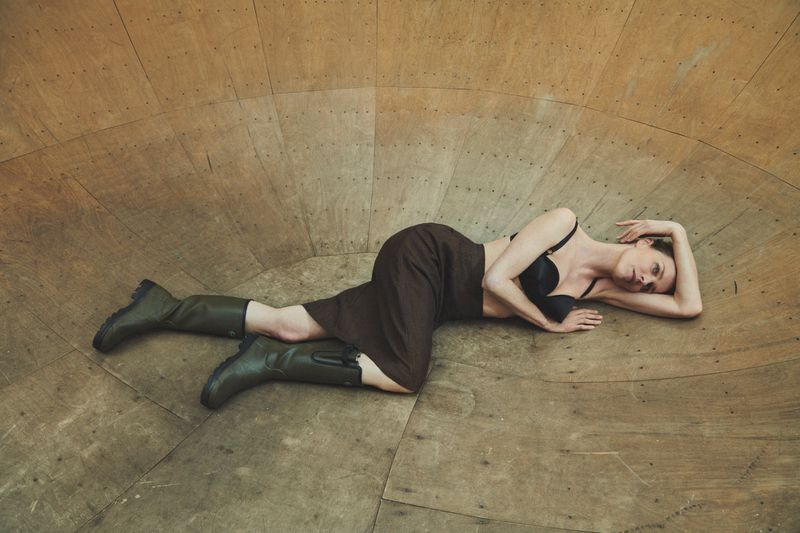 Woman in bra top, brown skirt, and green boots reclines in a rounded wooden structure, posing for a fashion editorial photo.