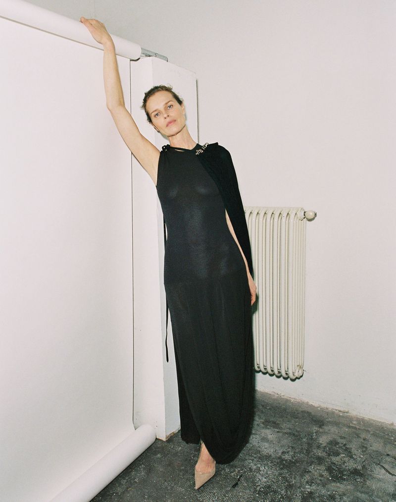 Adult woman in a long black dress leaning against a white wall with one arm raised, in a minimalist indoor setting with concrete floor.