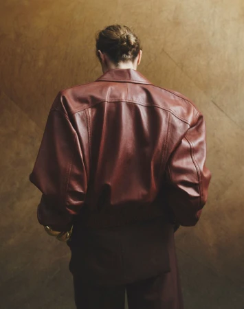 A person stands facing away wearing a red leather jacket and matching pants against a tan textured background with a gold bracelet.