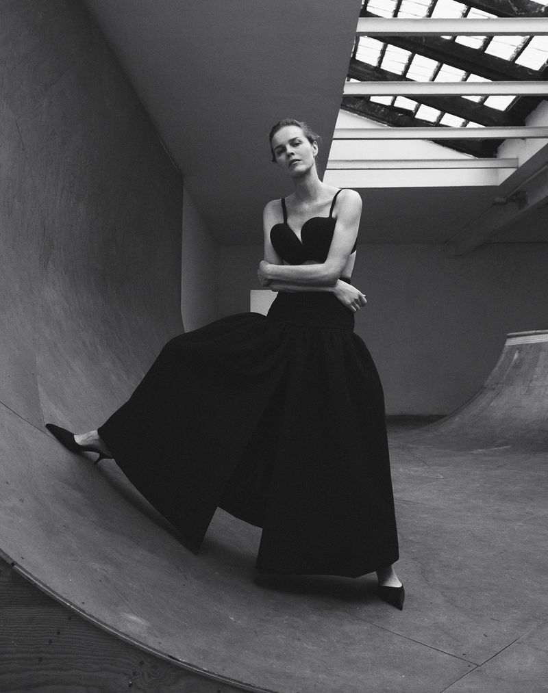 A woman in an elegant black dress poses on a concrete ramp in a black and white fashion photograph with dramatic natural lighting.