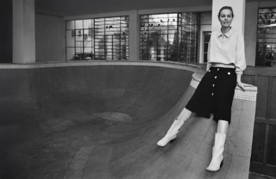 Eva Herzigova seated on the edge of an indoor skate ramp wearing a collared top, dark skirt, and light boots.