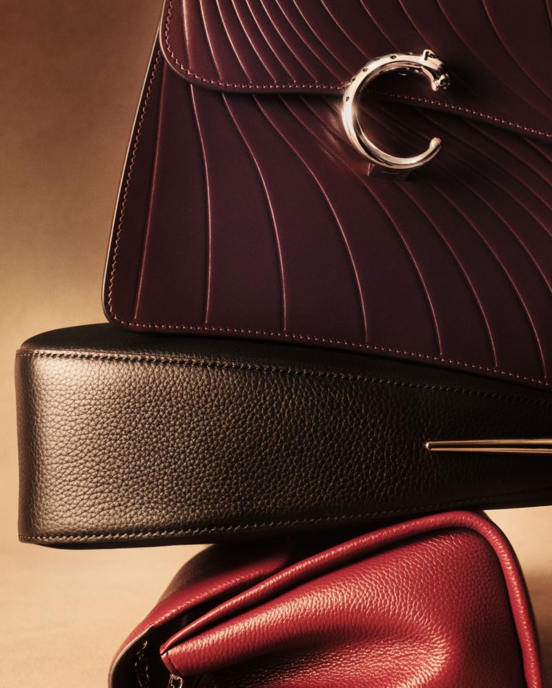 Close-up stack of three luxury leather handbags in burgundy, black, and red with silver hardware details against a neutral background.