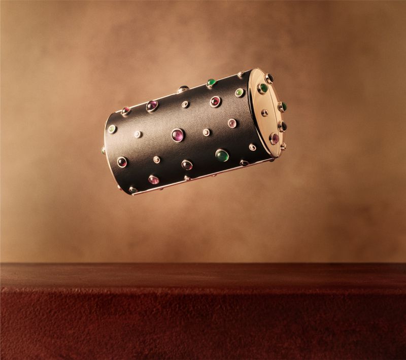 A cylindrical jeweled luxury case floats above a dark red wooden surface against a textured brown background.