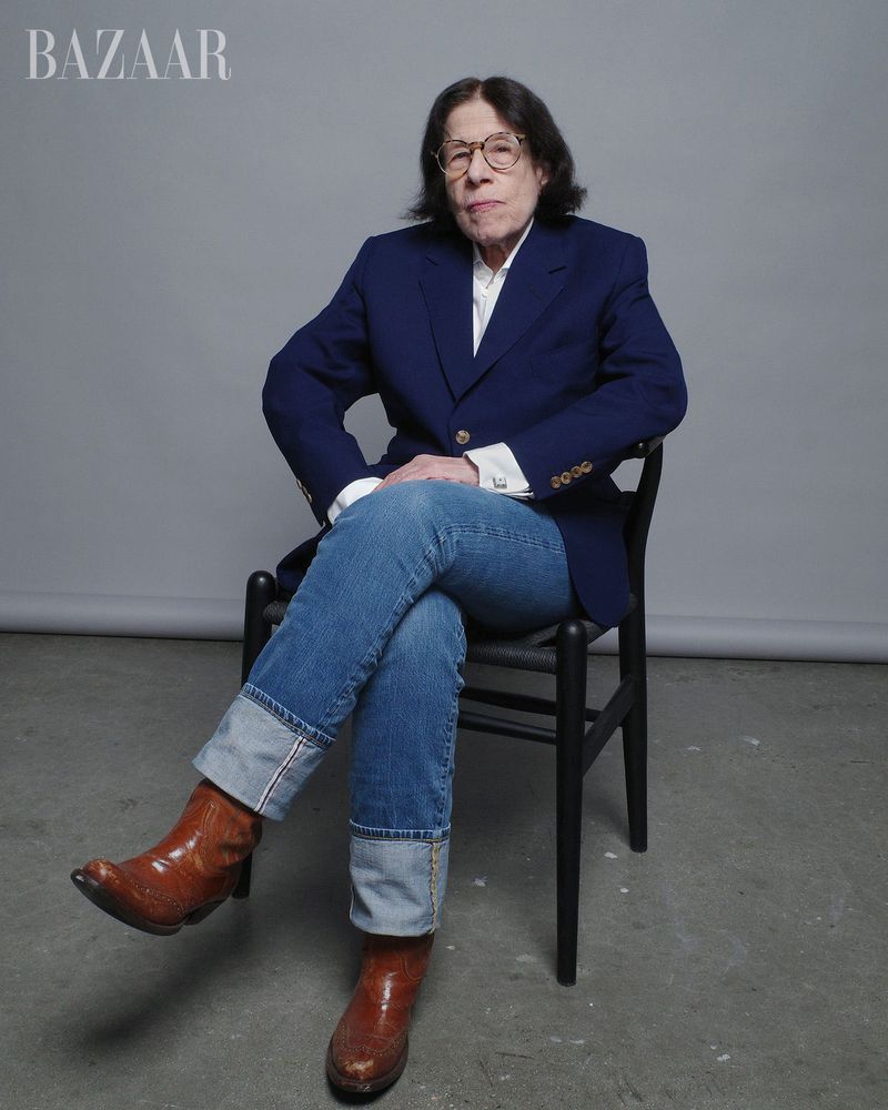 Portrait of an elderly woman with short brown hair wearing a blue blazer, jeans, and brown boots, seated on a black chair against gray backdrop.
