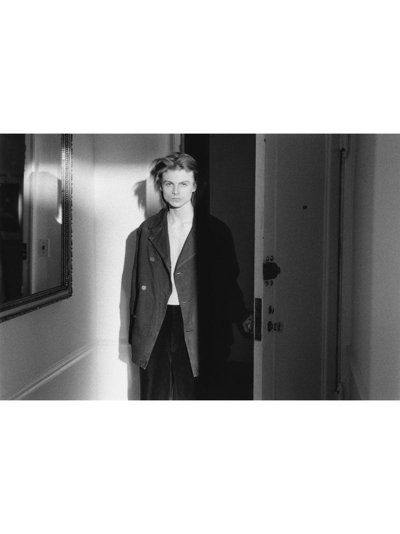 Monochrome portrait of a person standing in a doorway wearing a dark jacket inside an apartment hallway.