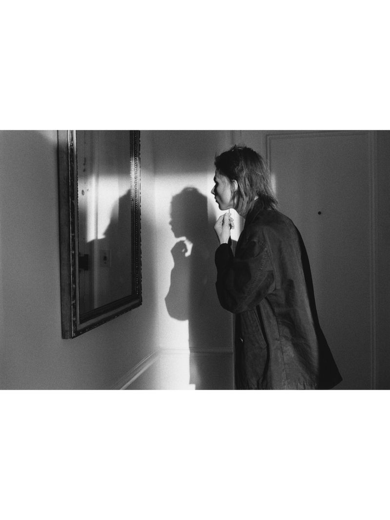 Monochrome image of a person standing in profile looking at a wall mirror with a strong shadow cast behind them.