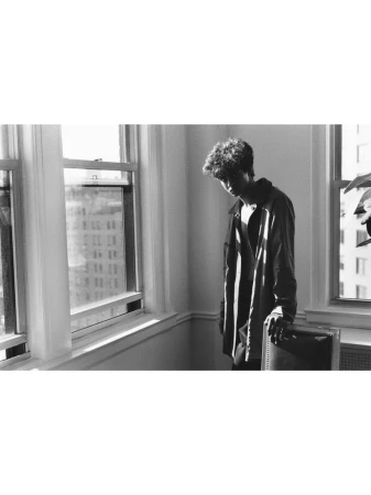 Monochrome portrait of a young man with curly hair standing near large windows in an interior room.