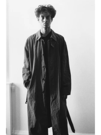 Monochrome portrait of a young man wearing a long dark coat and dark shirt against a simple indoor background.