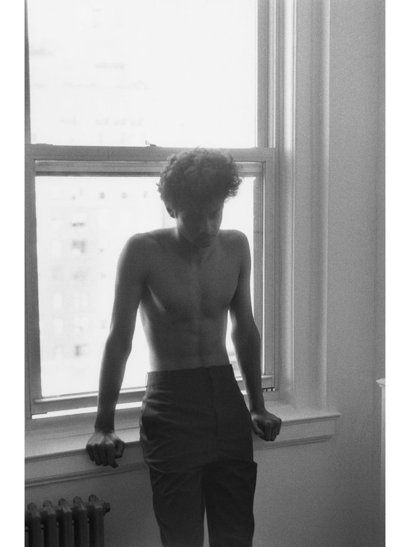 Monochrome portrait of a shirtless young man leaning against a window frame for Crosscurrent