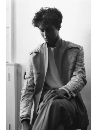 Portrait of a young man wearing a layered jacket seated indoors against a plain wall