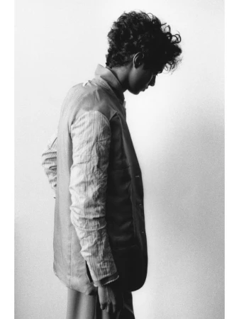 Monochrome portrait of a curly-haired man in profile wearing a collared jacket against a plain background